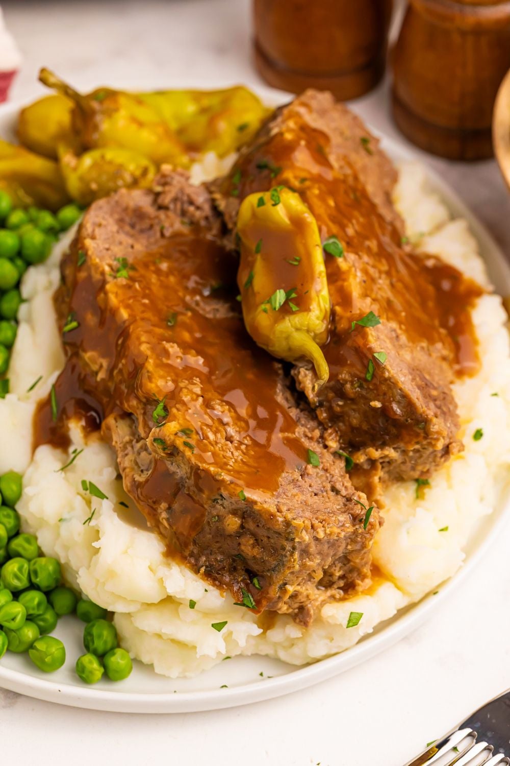 Crockpot Mississippi Meatloaf sliced and placed on mashed potatoes with some peas to side and wooden salt and pepper shakers