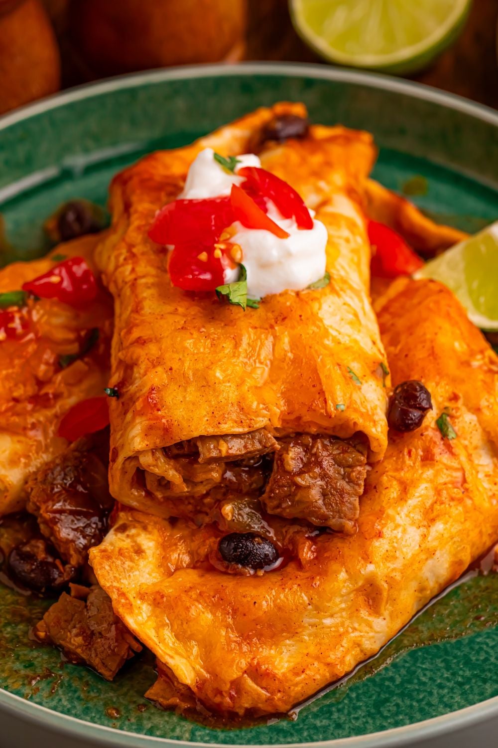 Steak Enchiladas on a plate with some sour cream and salsa on top with a few limes to side of the plate on wooden table