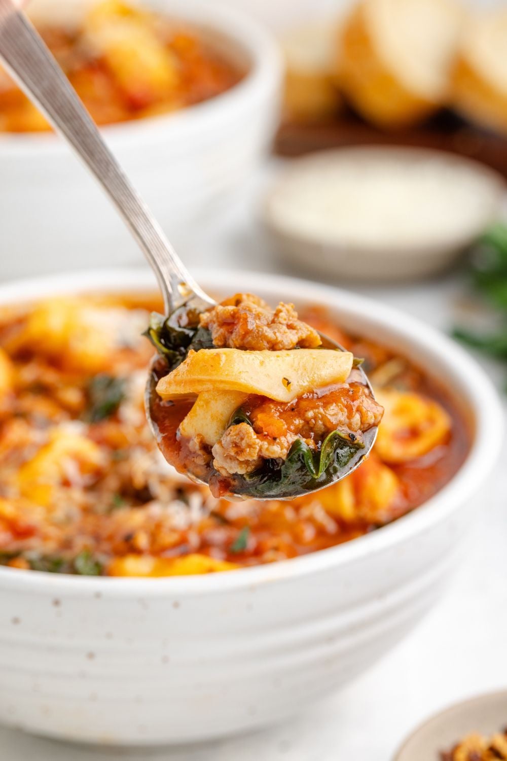 a white bowl that has Sausage Tortellini Soup in it with a spoon with some of the soup above the bowl to show a closer look at the soup
