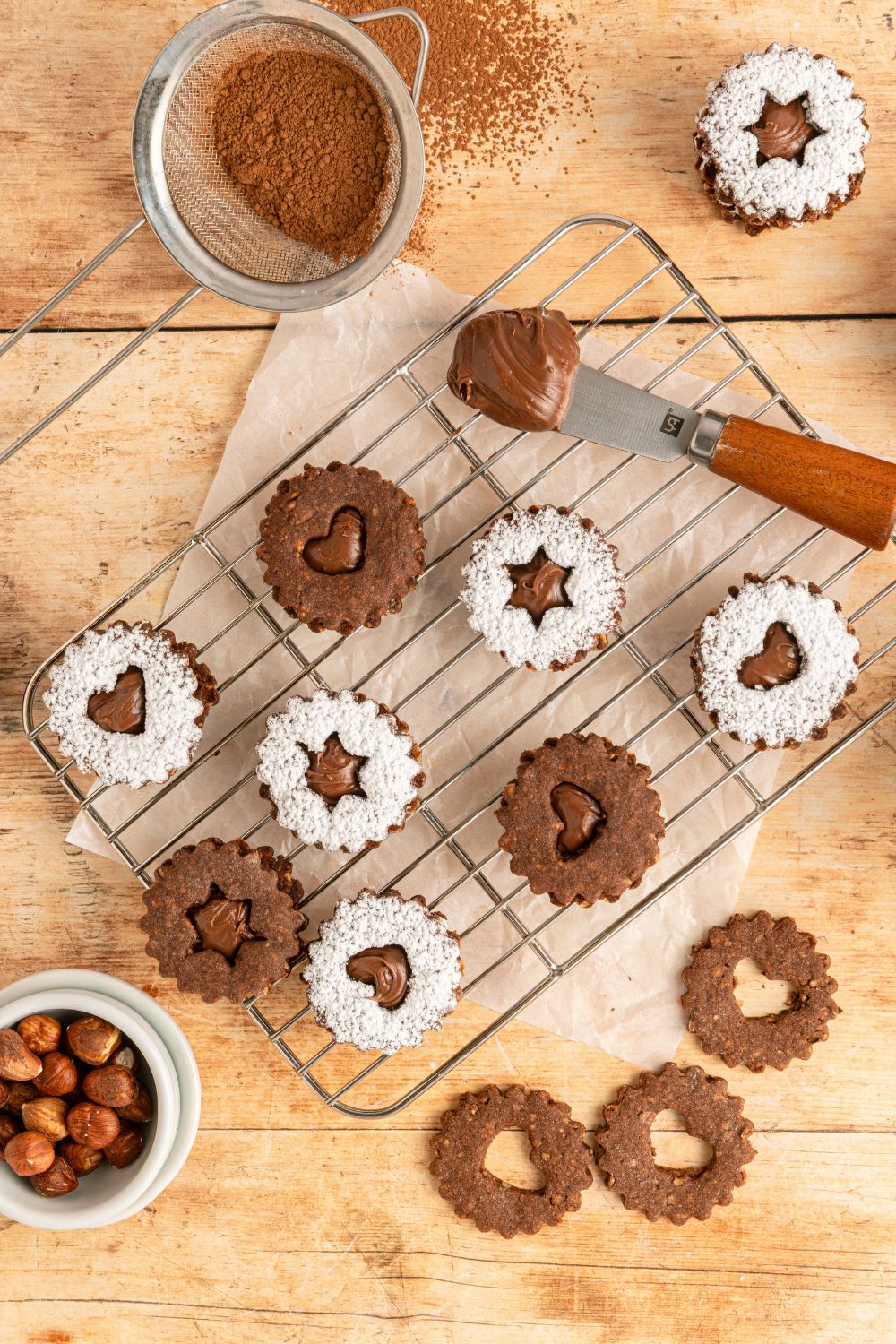 Chocolate Linzer Cookies on a table with a cooling rack with some cookies assembled and others just placed around the table 
