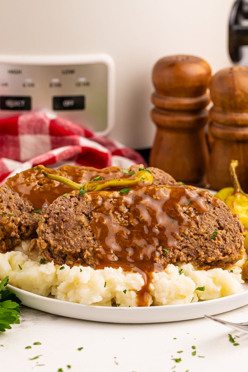view of table with mashed potatoes with slices of meatloaf on a plate with a red checkered towel with slow cooker in the background