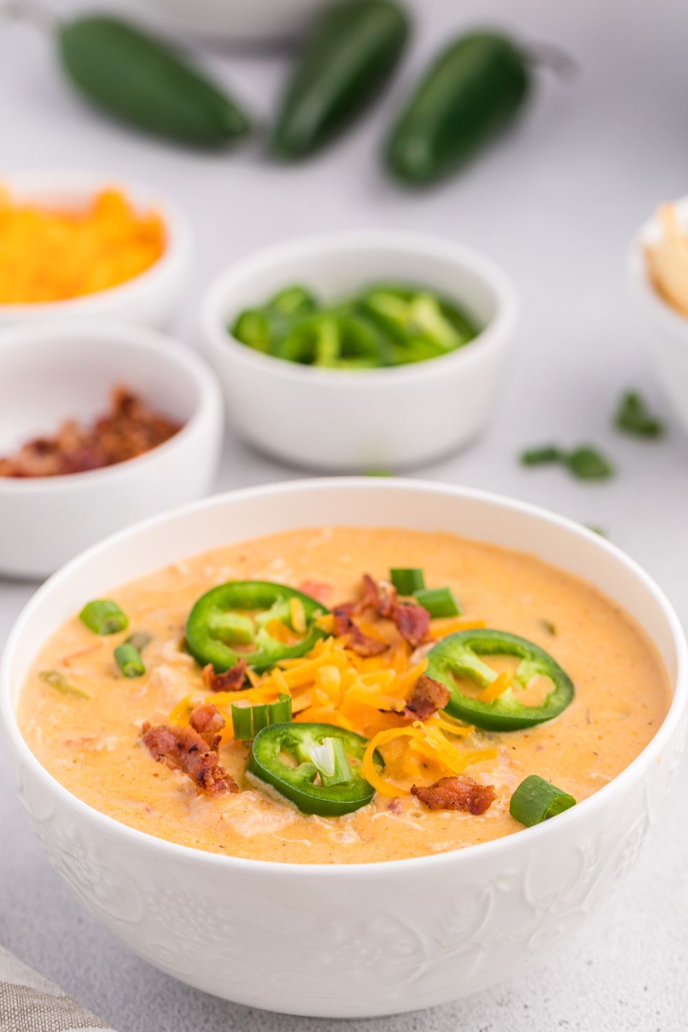 jalapeño popper crack chicken soup in a white bowl with more of the ingredients around the table and whole jalapenos behind it on table