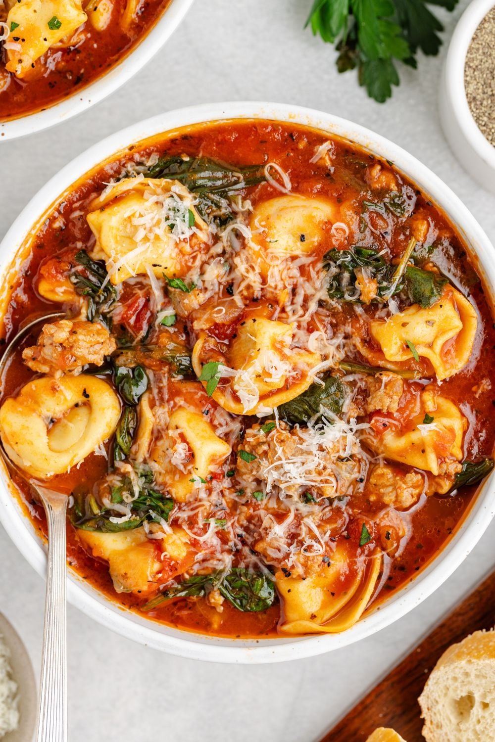 a white bowl full of Sausage Tortellini Soup on a white table with a spoon on side of bowl