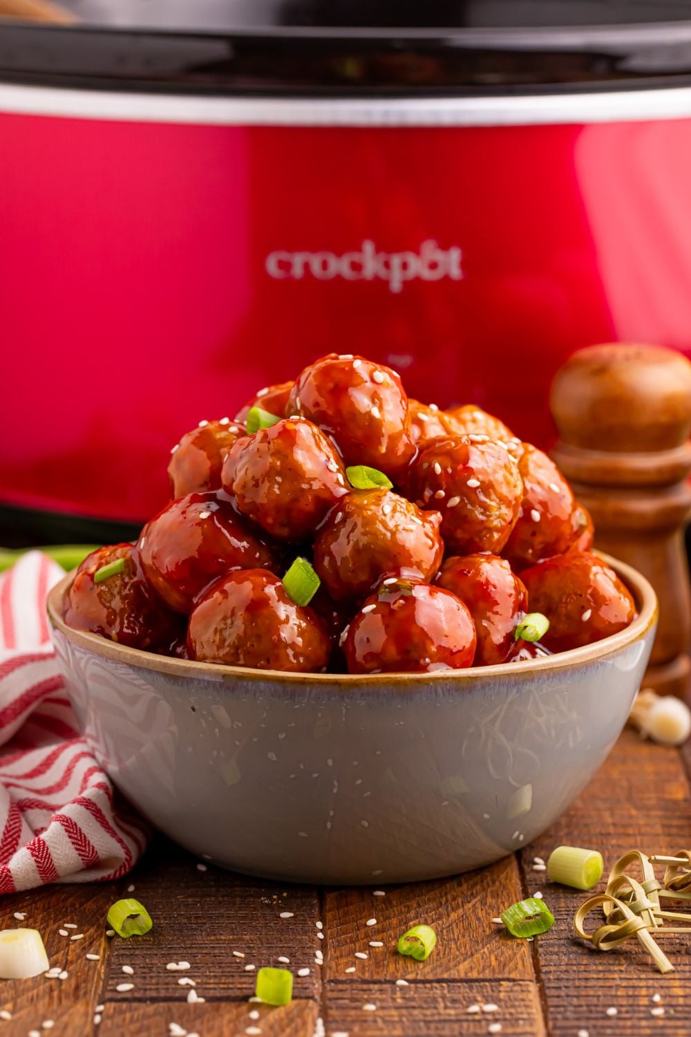 Crockpot Sweet Chili Meatballs in a bowl in front of a crockpot on table with green onions spread around the table and a red and white towel