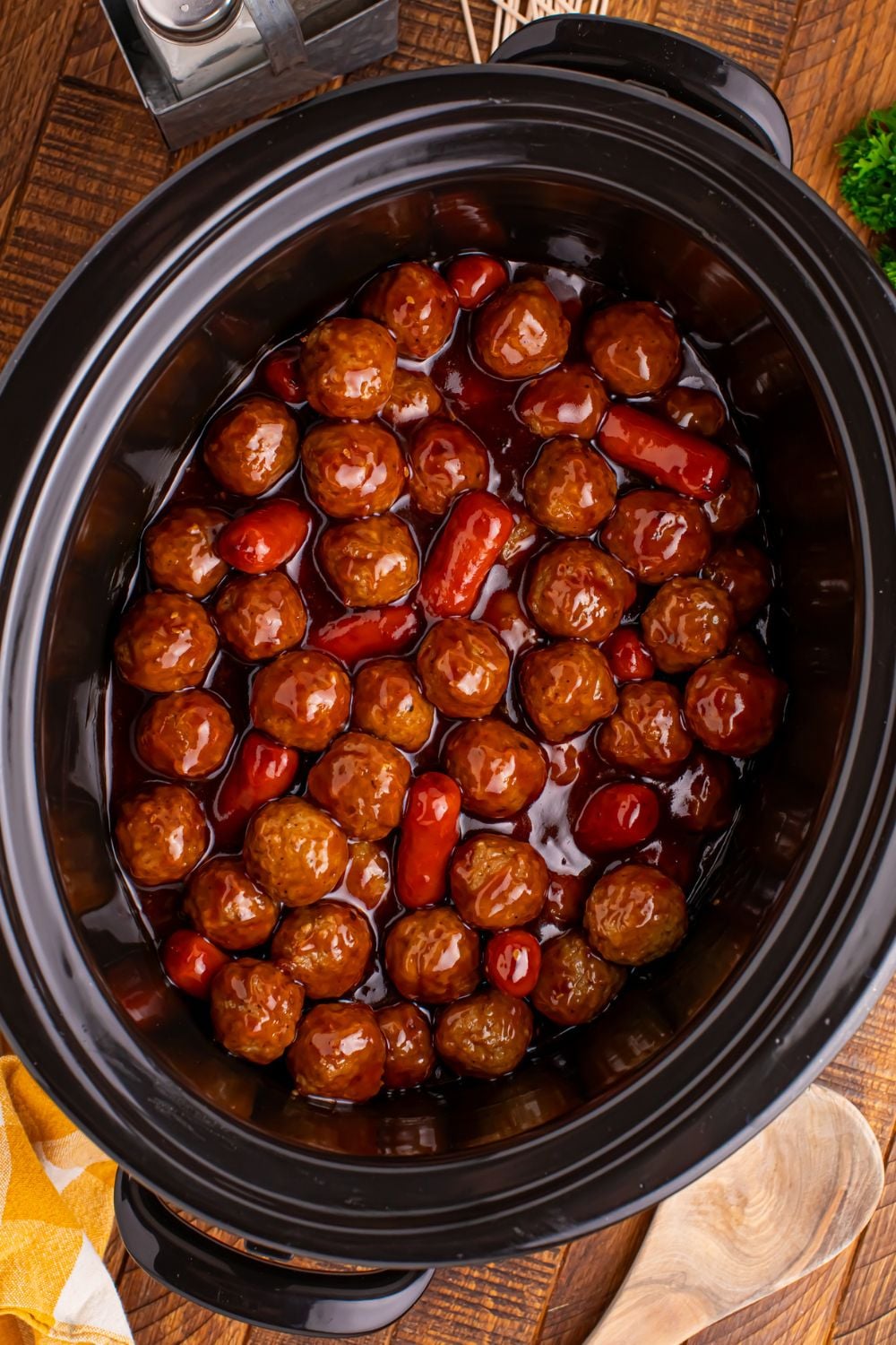 a black Crockpot full of Teriyaki BBQ Cocktail Meatballs & Lil Smokies with a wooden spoon to side on table and salt and pepper shakers 