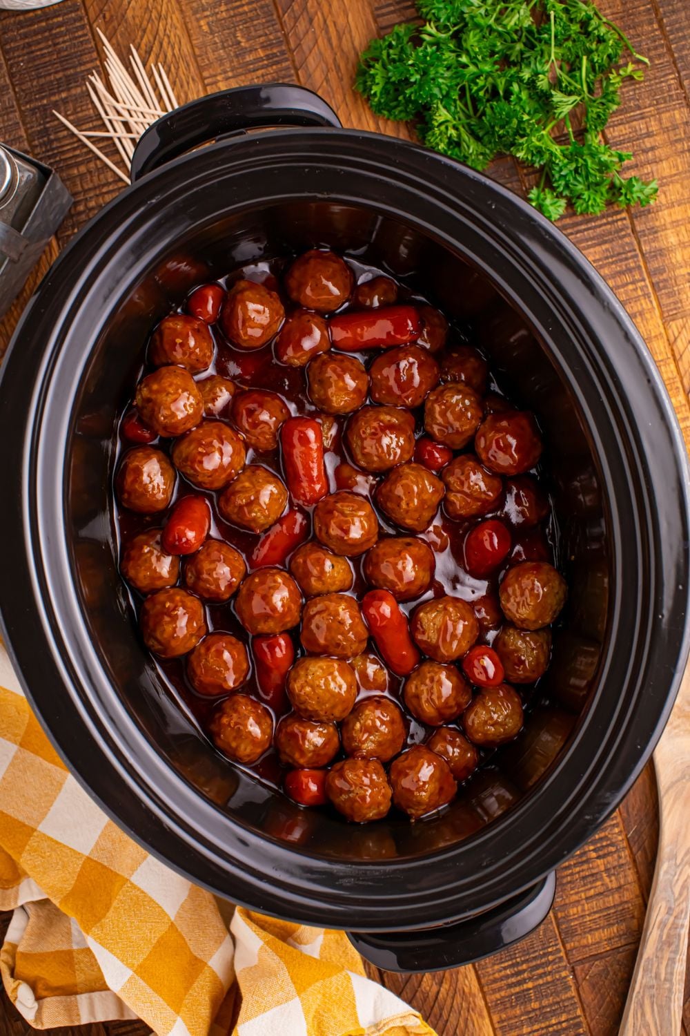 a tilted black crockpot on a wooden table full of smokies and meatballs in sauce 