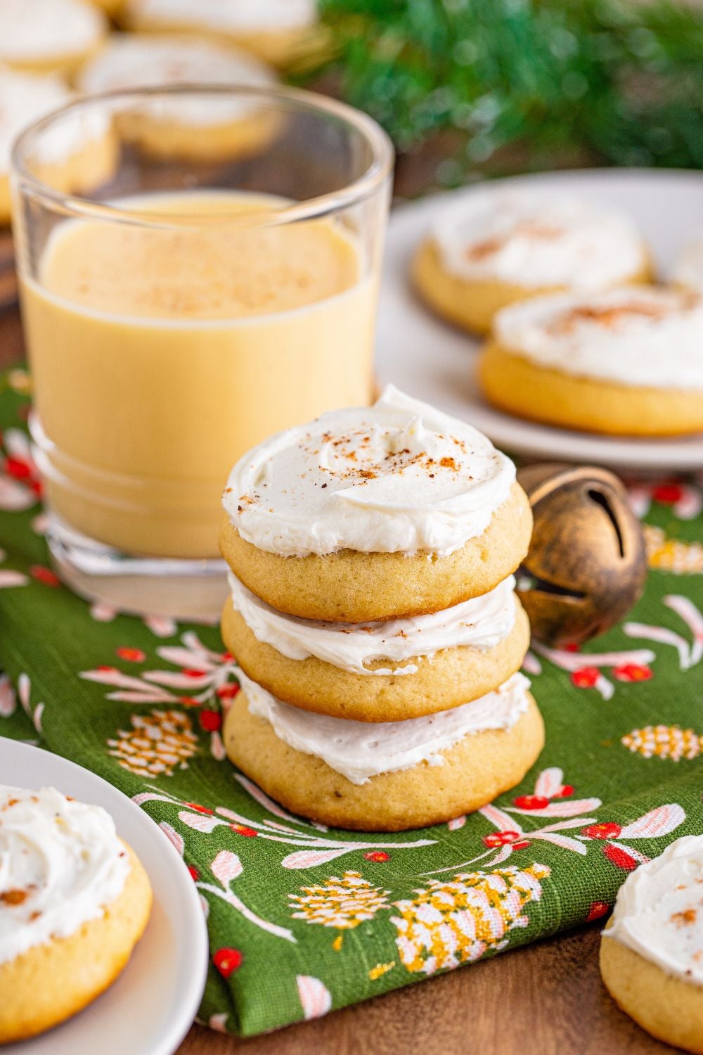 Soft Frosted Eggnog Cookies stacked on a table with more plates of cookies around and a glass full of eggnog
