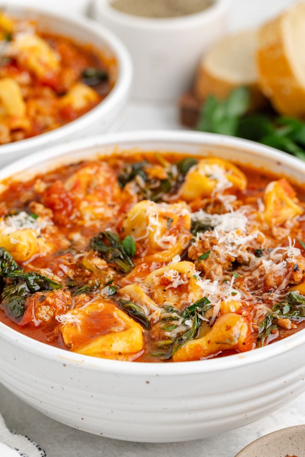side view of a white bowl full of Sausage Tortellini Soup with parmesan cheese and some parsley to side on table