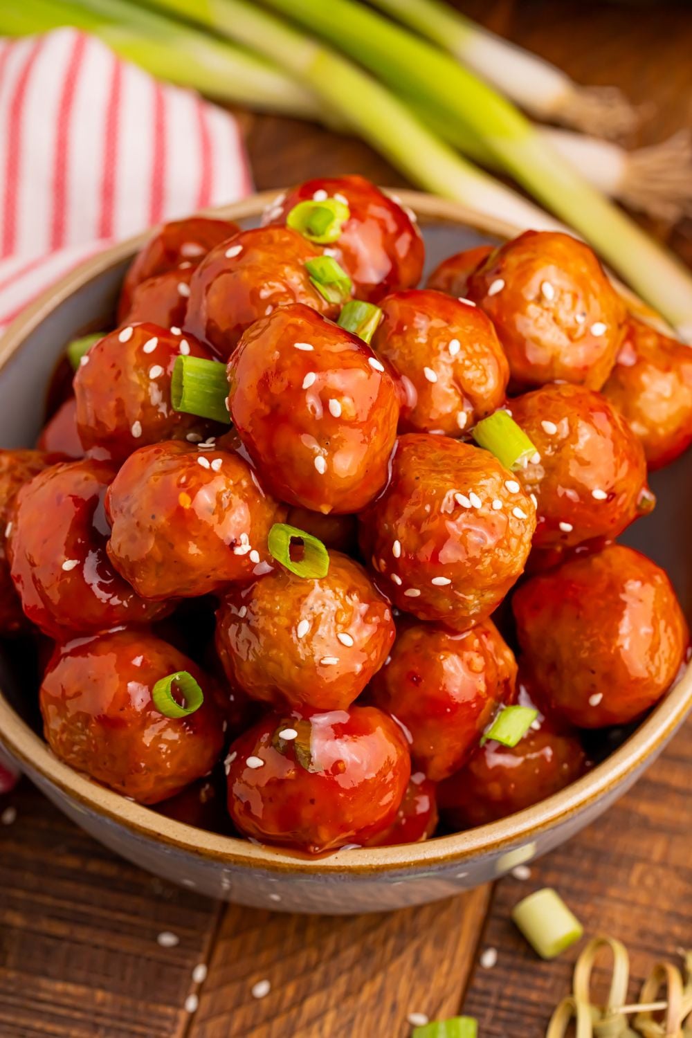 Crockpot Sweet Chili Meatballs in a wooden bowl on table with sesame seeds and green onions sprinkled on top with more onions behind it on the table