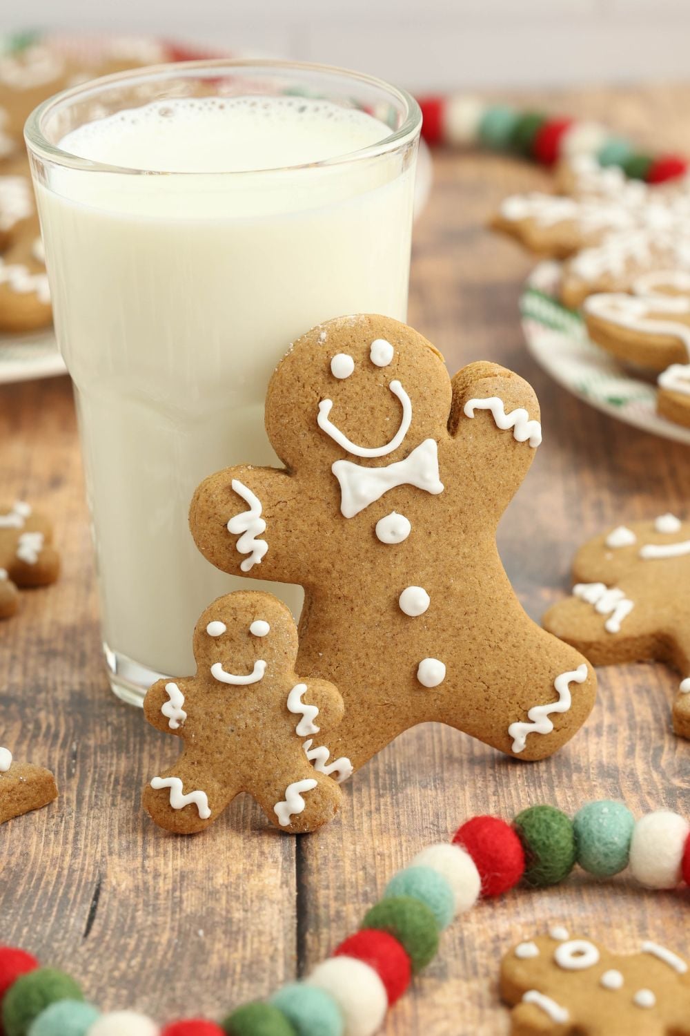 tall glass of milk with two gingerbread cookies standing on side and more cookies around the table 