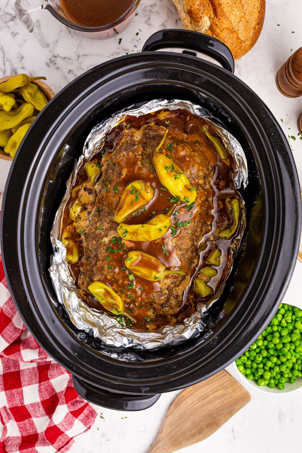 black crockpot with Mississippi Meatloaf with gravy and peppers on top and peas and peppers to the side