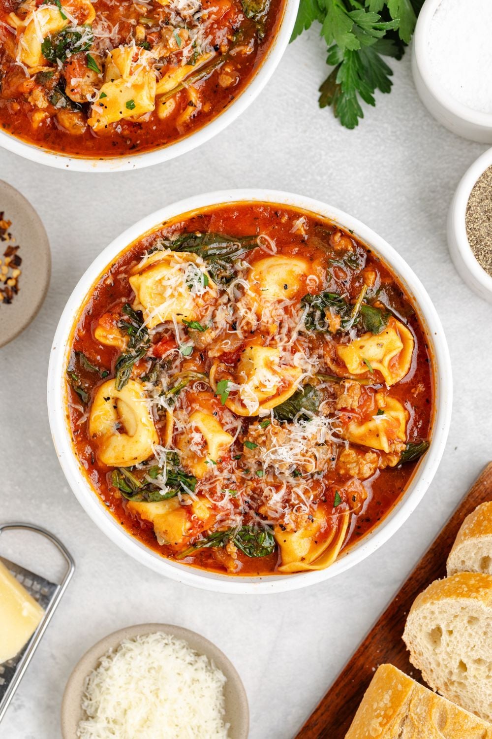a white table with white bowls full of Sausage Tortellini Soup and topped with parmesan cheese, and grated cheese in a small bowl, and sliced French bread to side to create a dinner themed table