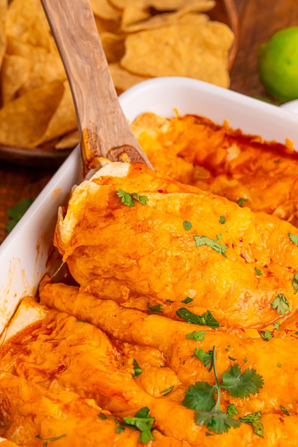white enchilada pan with a wooden spatula scooping up one of the enchiladas from the pan with tortilla chips around the table