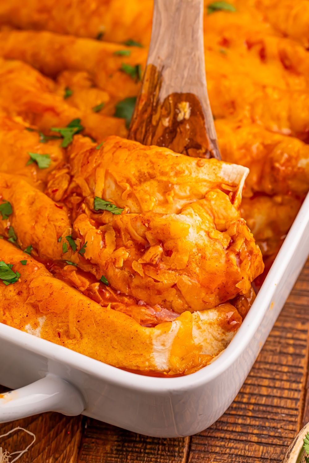 Steak Enchiladas in a white casserole dish with a wooden spoon scooping up an enchilada our of the pan