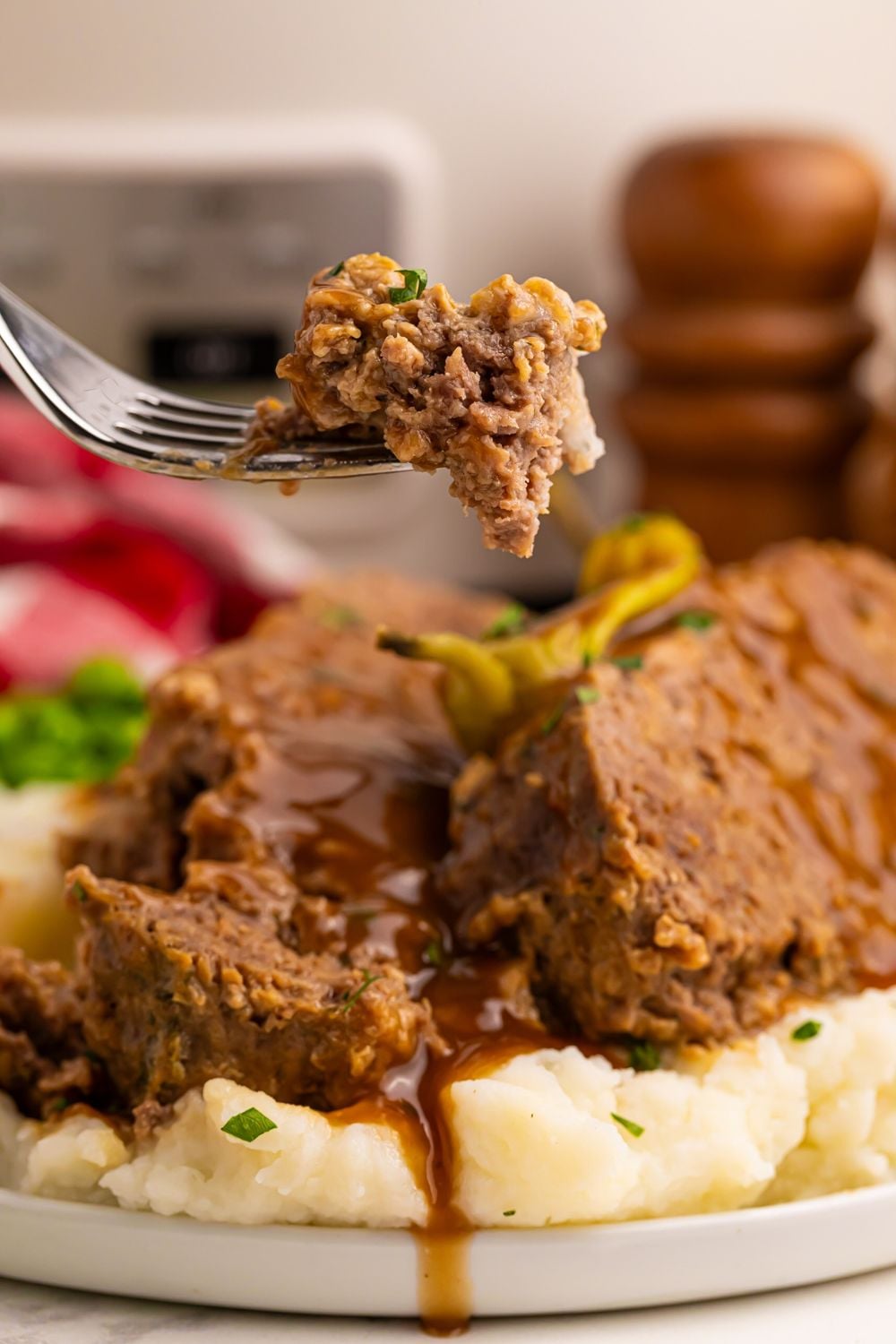 Crockpot Mississippi Meatloaf on a plate with a fork with a bite on it with a plate under full of more meatloaf and mashed potatoes