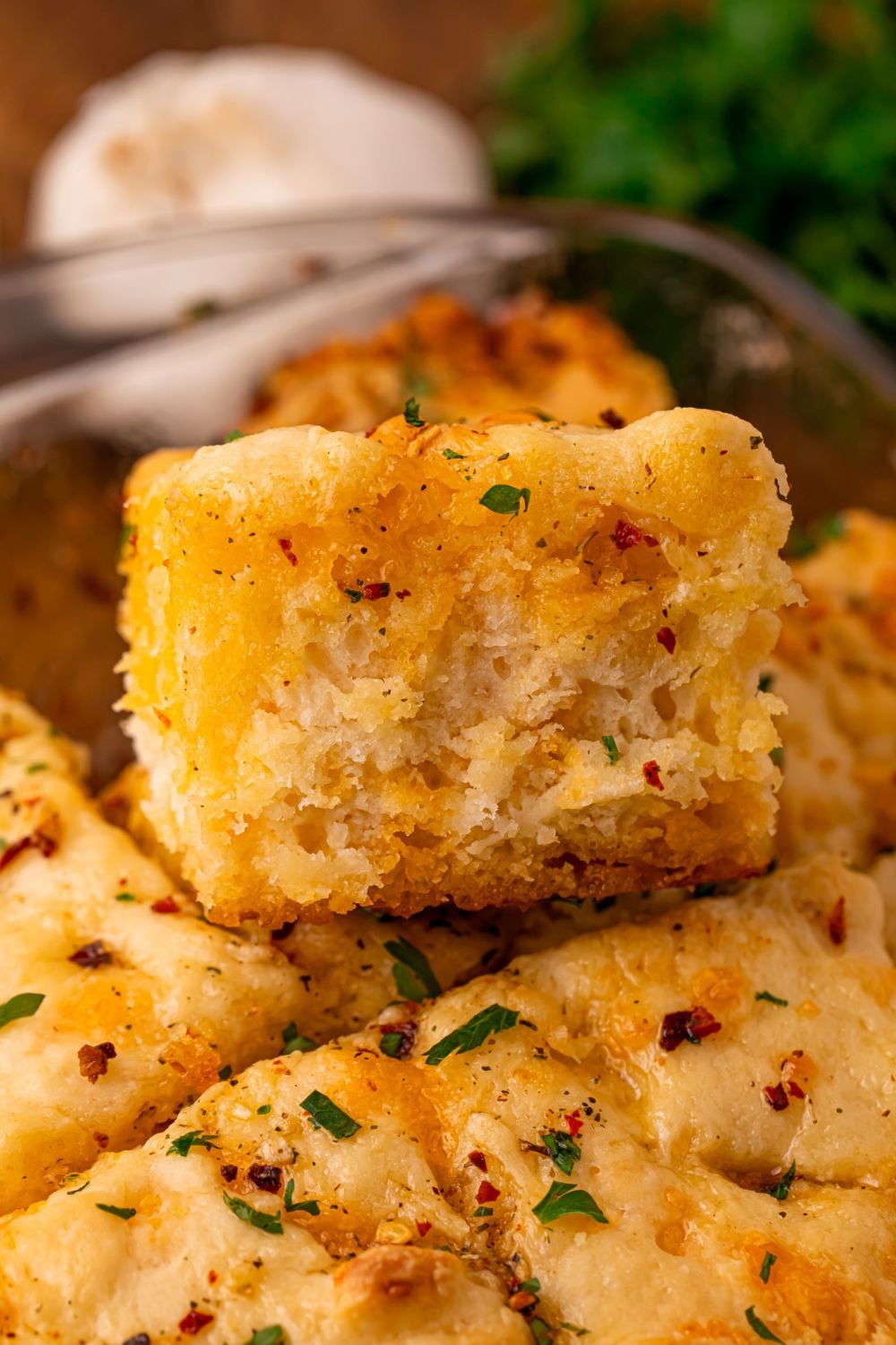 Cowboy Butter Swim Biscuits in a glass pan with one of the biscuits being lifted out of the pan a bit with parsley behind it on table and a bulb of garlic