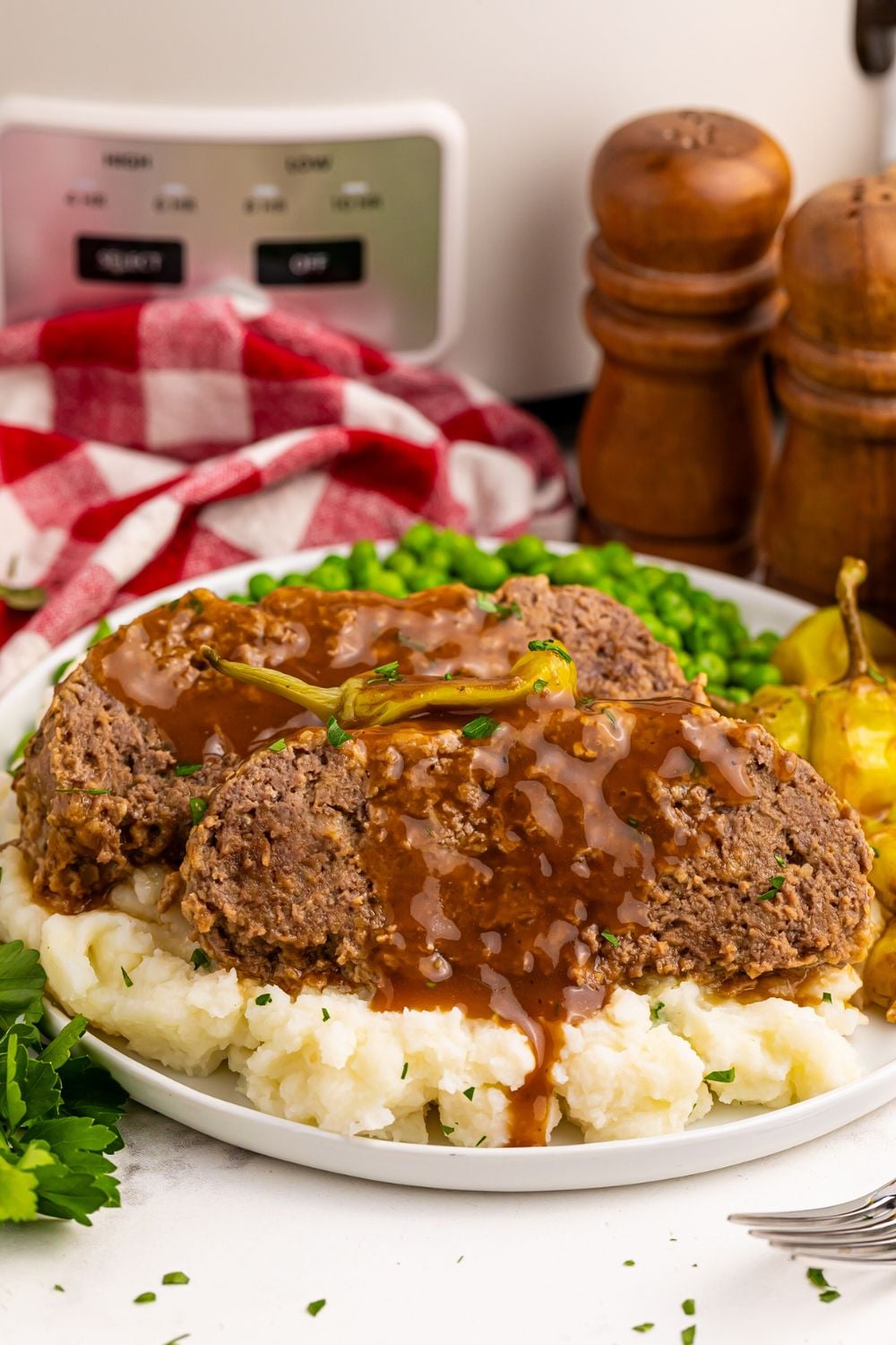 a table set up for a meal with a plate full of mashed potatoes, Mississippi meatloaf, peas and salt and pepper shakers behind the plate and a crockpot
