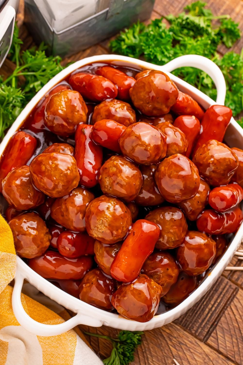 looking down on table to see a container full of Crockpot Teriyaki BBQ Cocktail Meatballs & Lil Smokies with some fresh parsley around the table and a yellow towel to the side 