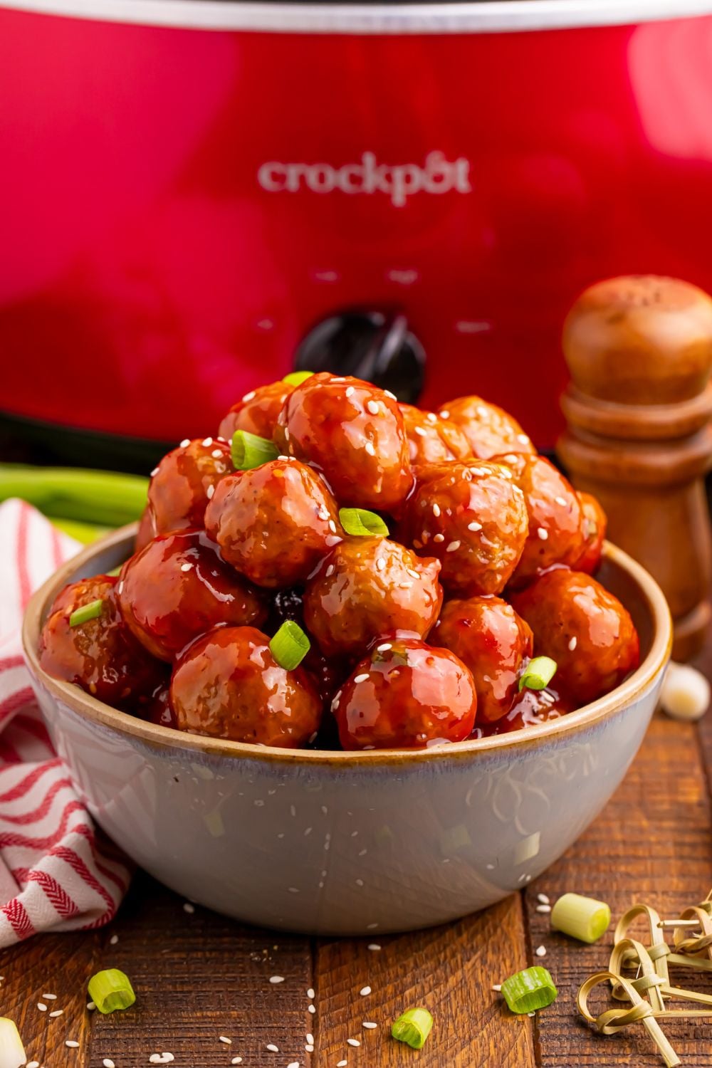 a red crockpot sitting on table with a bowl of meatballs and chopped green onions sprinkled on top and around the table with a salt shaker by the crockpot