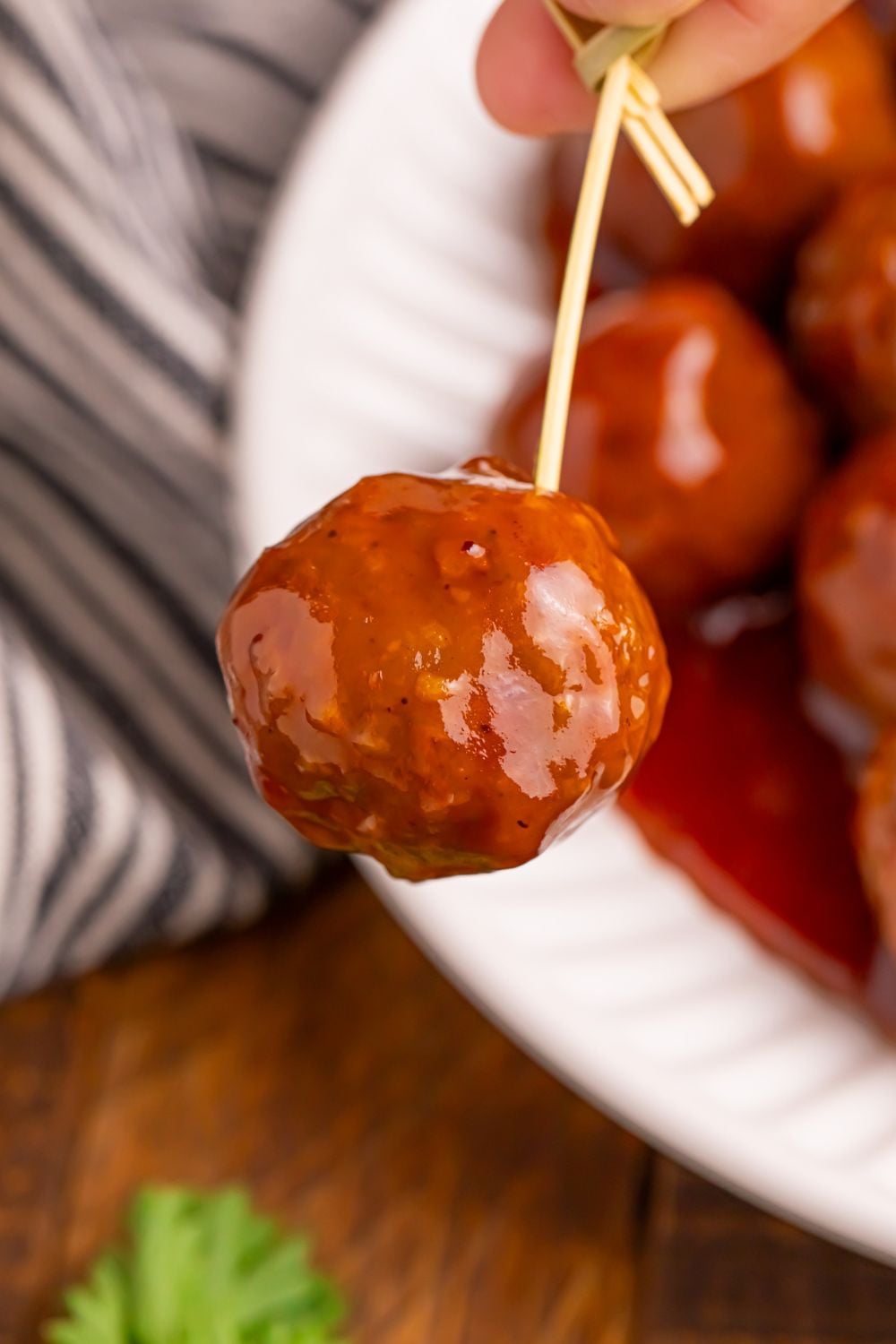 toothpick being held with a Peach BBQ Meatball on it and a plate full of more meatballs below on table