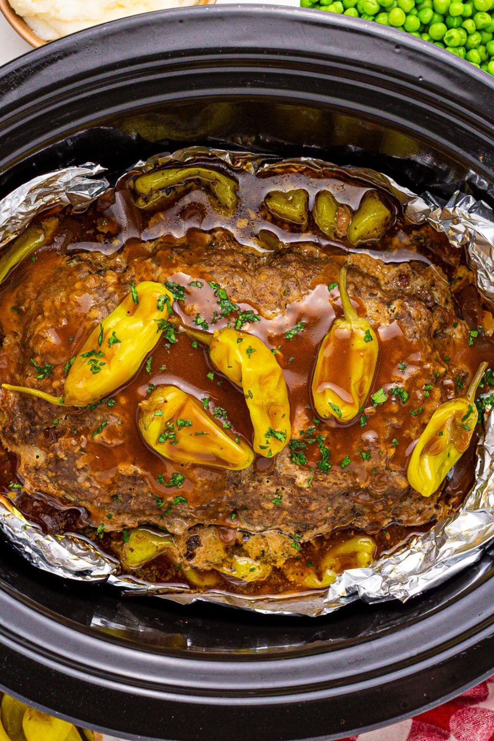 black slow cooker side ways on the table with meatloaf cooked in the slow cooker with gravy and peppers on top