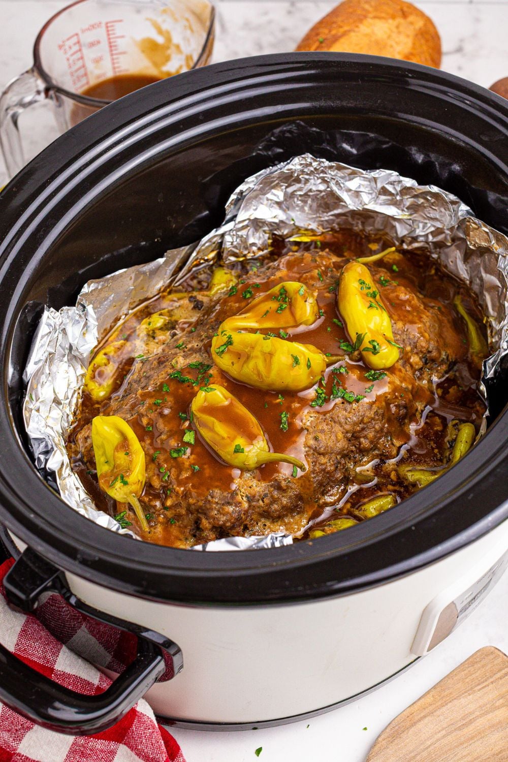 crockpot on table with foil and inside is a meatloaf with gravy and peppers on top with a measuring cup with gravy behind it on table and an onion