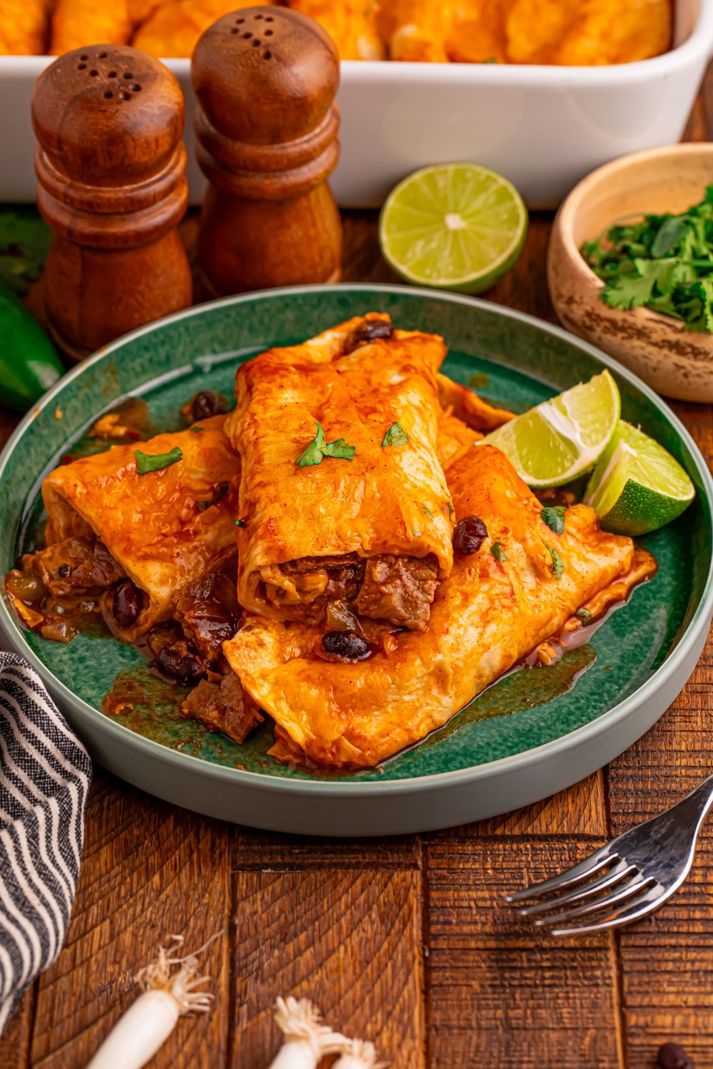 three Steak Enchiladas on a green plate that is sitting on a dinner table with a fork, salt and pepper shaker, bowl of cilantro and more around the table
