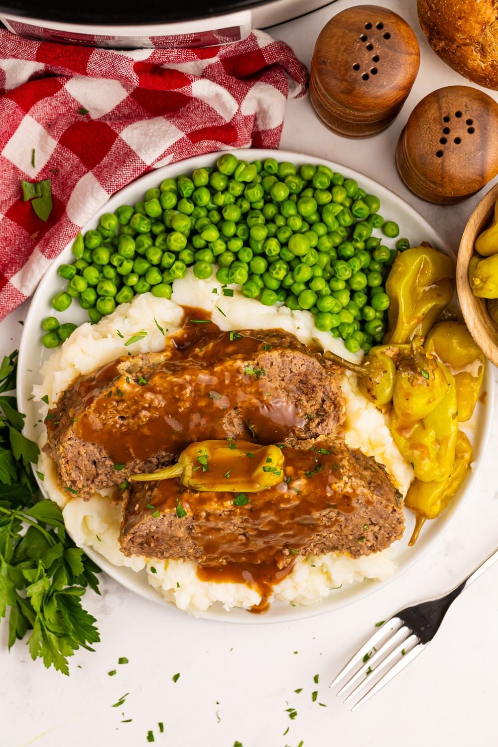looking down on table to a plate full of Crockpot Mississippi Meatloaf with mashed potatoes, peas, and peppers to the side with a red checkered towel in the background