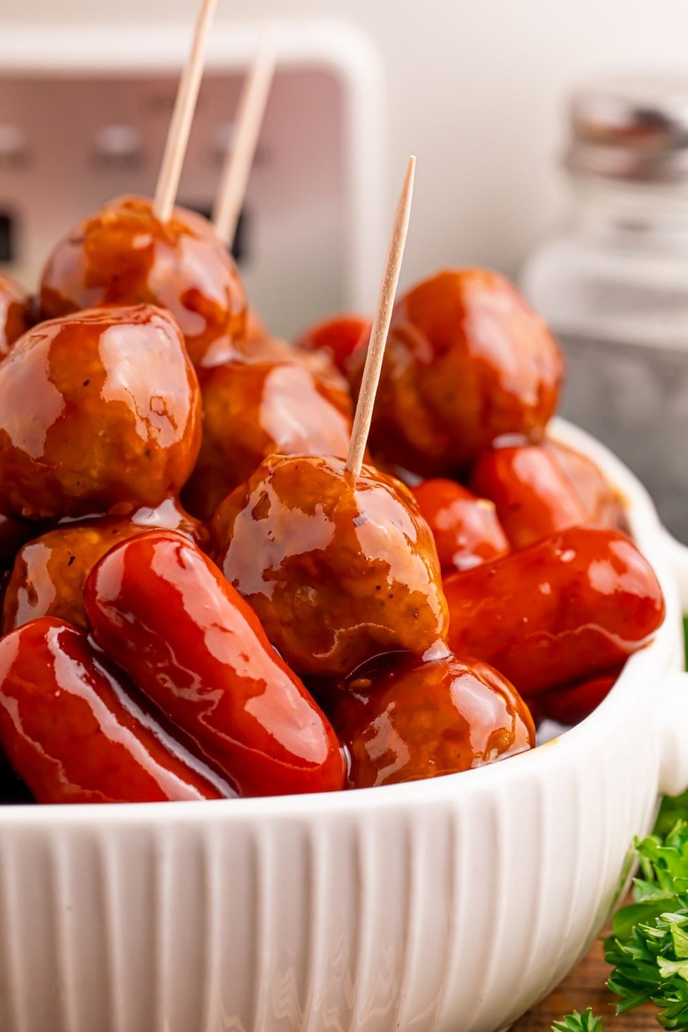 Crockpot Teriyaki BBQ Cocktail Meatballs & Lil Smokies in a white bowl on table with toothpicks poked into a few of them with the silver crockpot behind it on the table 