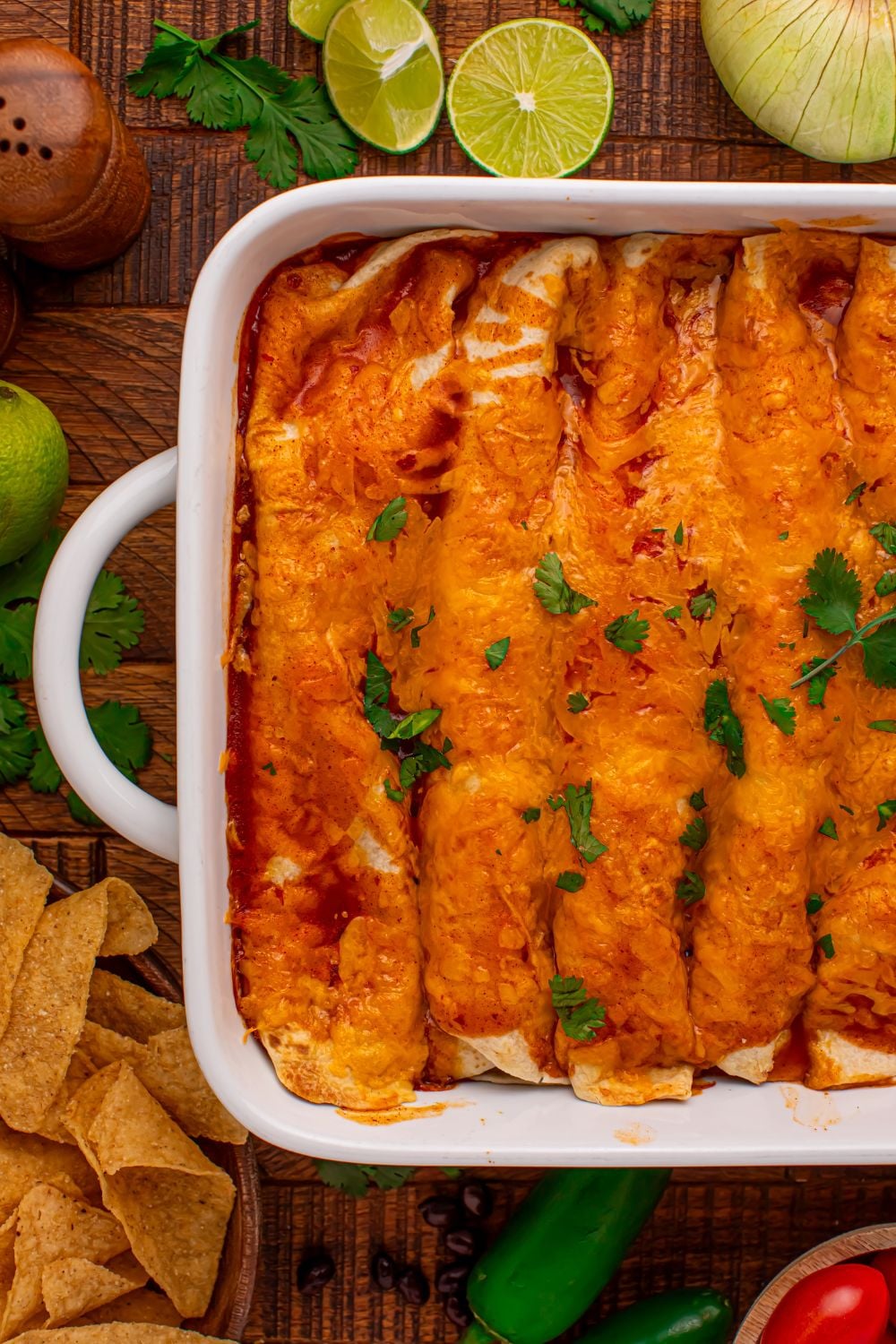 Steak Enchiladas in a white casserole dish with sliced limes on the wooden table and tortilla chips around the table