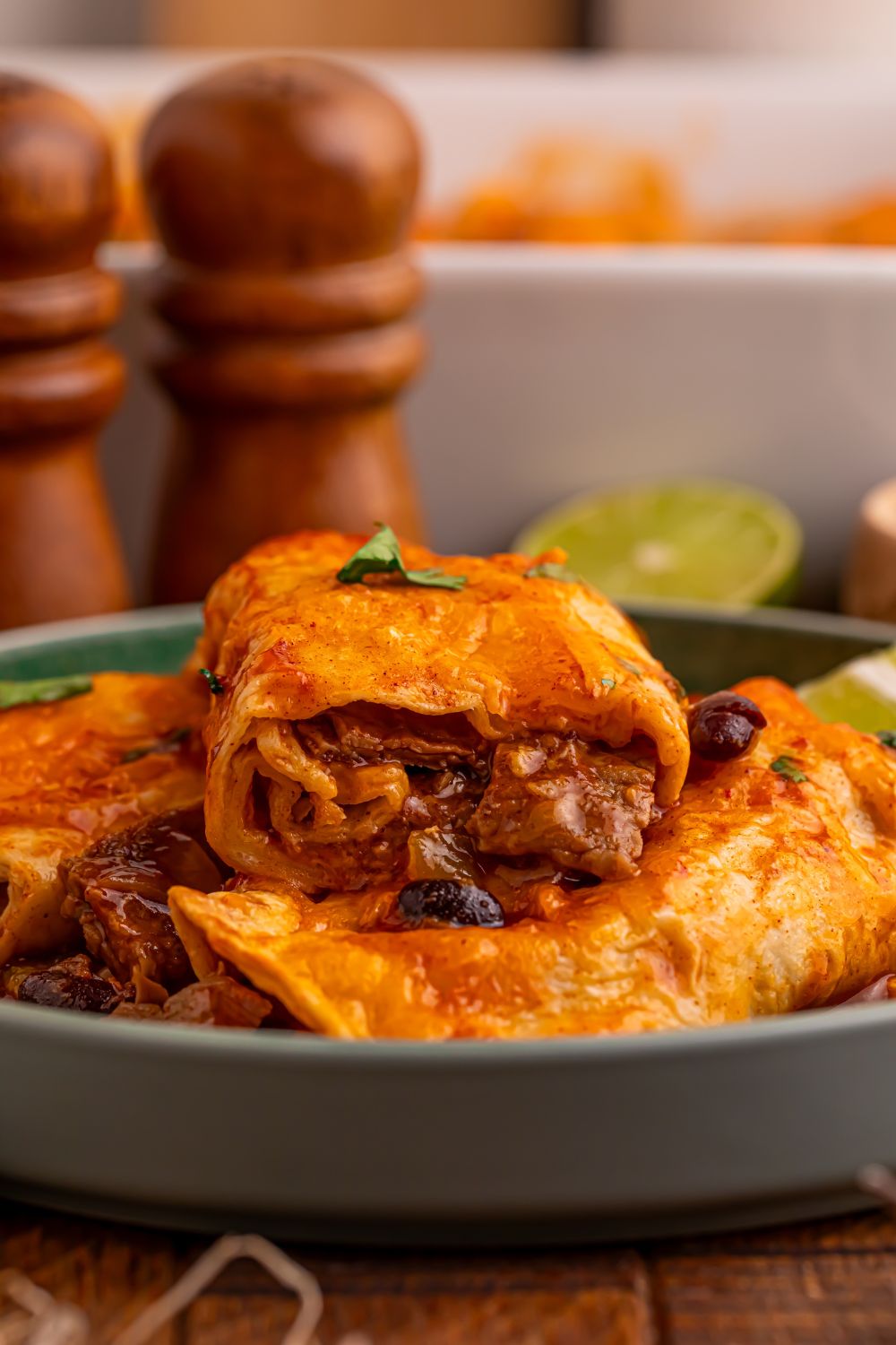 stack of Steak Enchiladas on a grey plate on table with some limes to the side and a white casserole dish behind it on the table