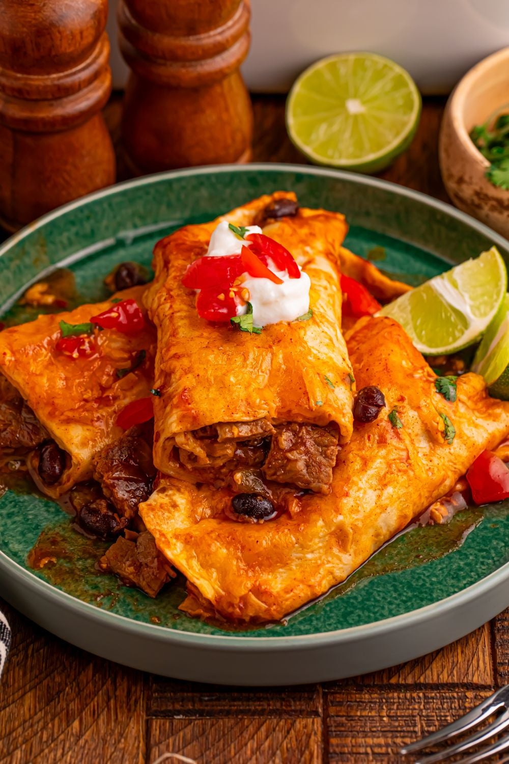 green plate with a few Steak Enchiladas on it and topped with sour cream, salsa, and lime wedge to side