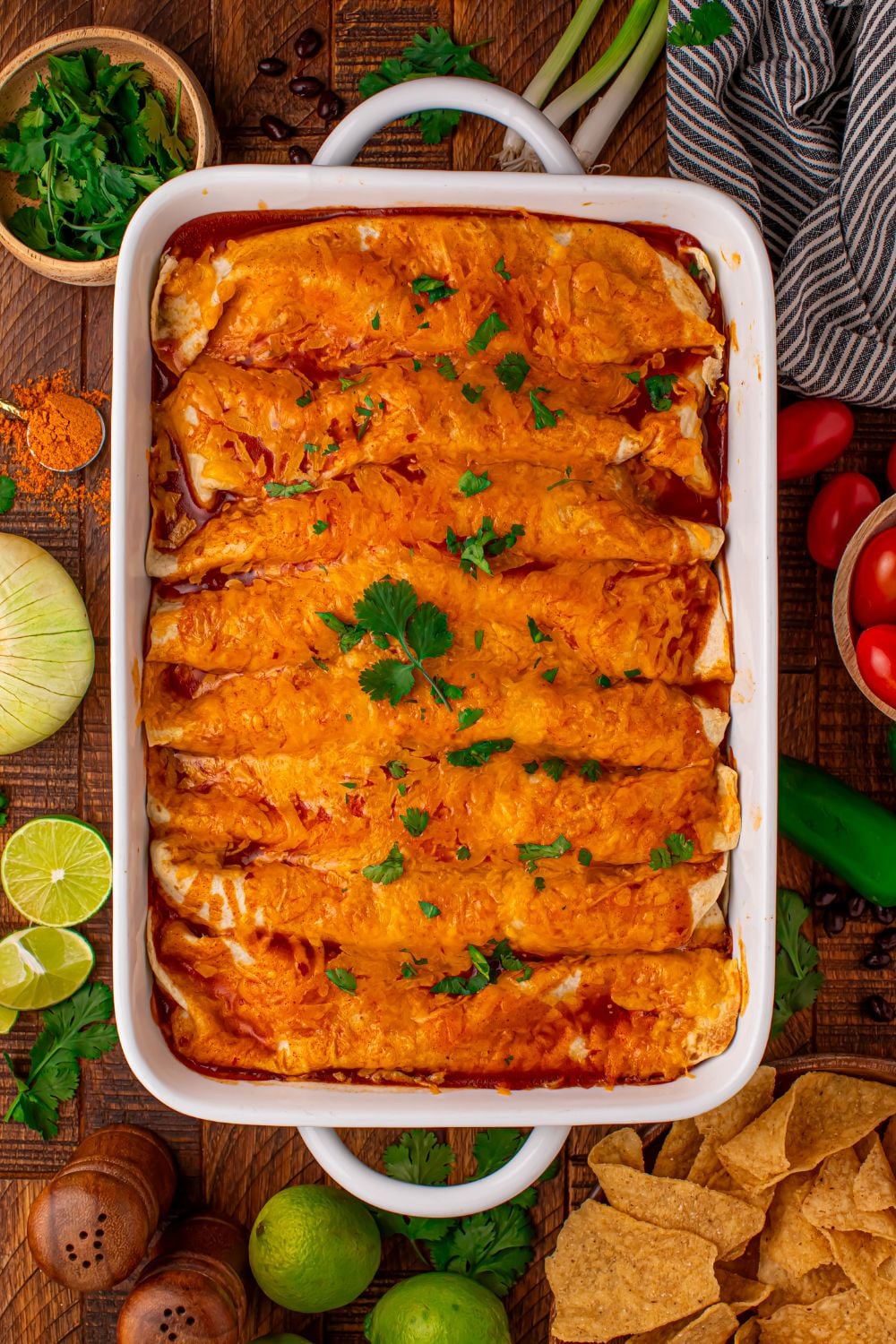 white 9x13 casserole dish on wooden table full of Steak Enchiladas with taco toppings around the pan