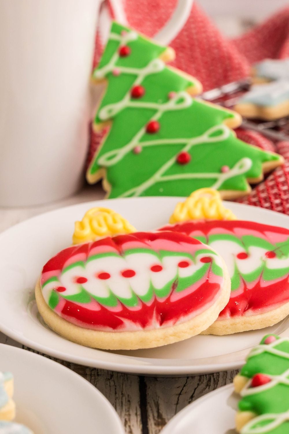 Tall glass of milk on table with Decorated Sugar Cookies around table and a plate with two of the cookies that look like Christmas ornaments