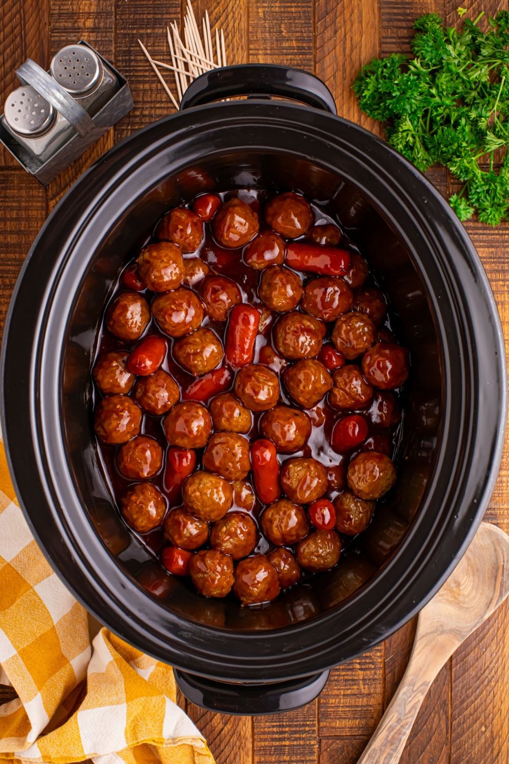 looking down on table to see a black crockpot full of Crockpot Teriyaki BBQ Cocktail Meatballs & Lil Smokies with salt and pepper shaker to side and a wooden spoon in front 