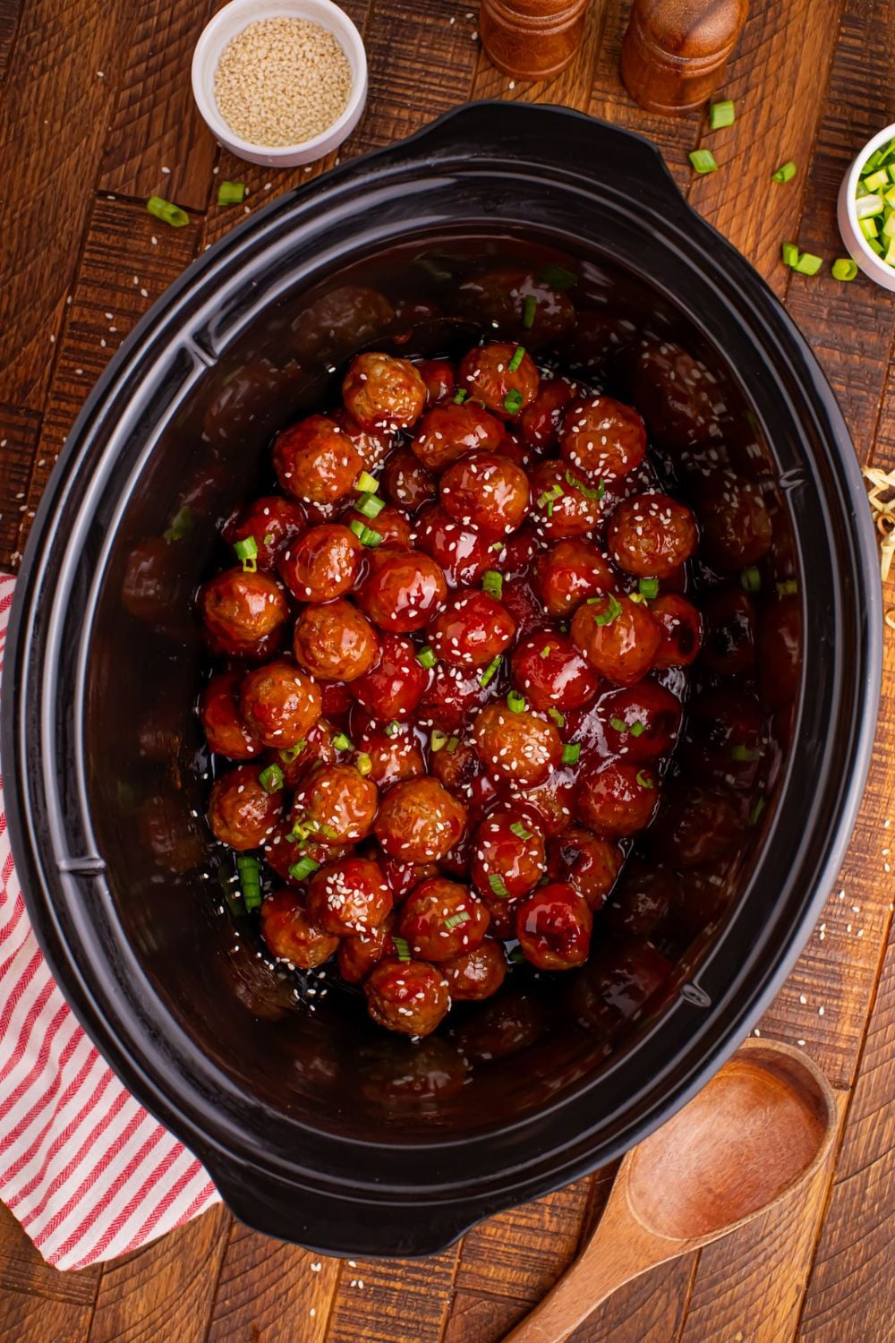 Crockpot Sweet Chili Meatballs in a black crockpot on table with a wooden spoon and a red striped towel to side