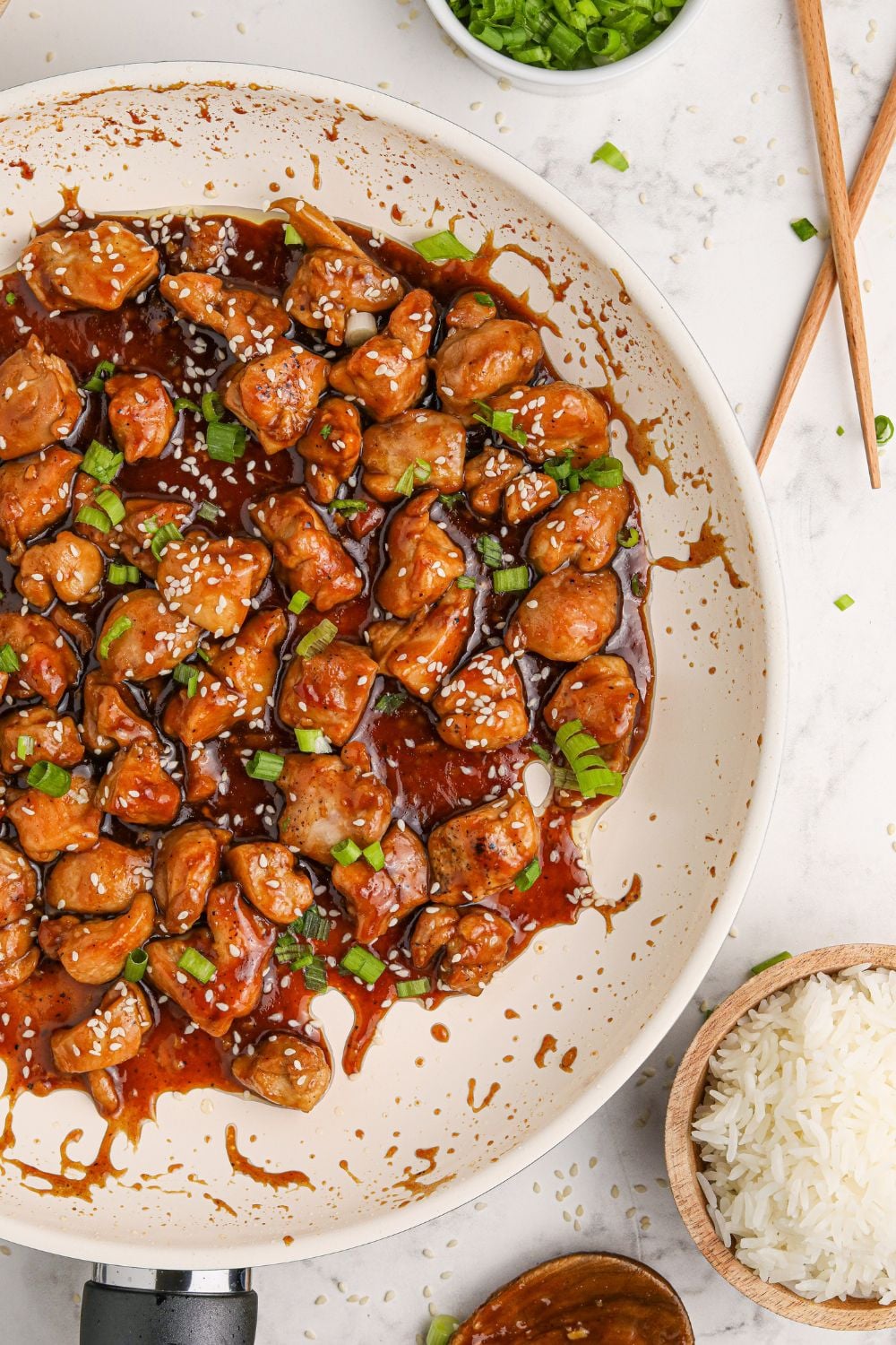 a white skillet full of saucy bourbon chicken on a table with chopsticks to the side and a bowl of rice to the side of the pan