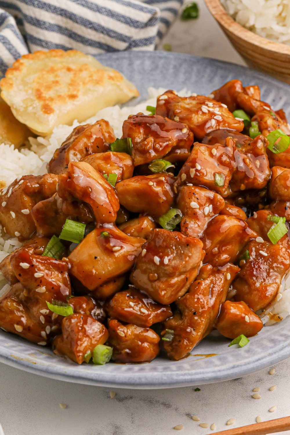 close up look of all the bourbon chicken on a blue plate on the table with some rice to the side of the plate and a bowl of more rice behind the plate