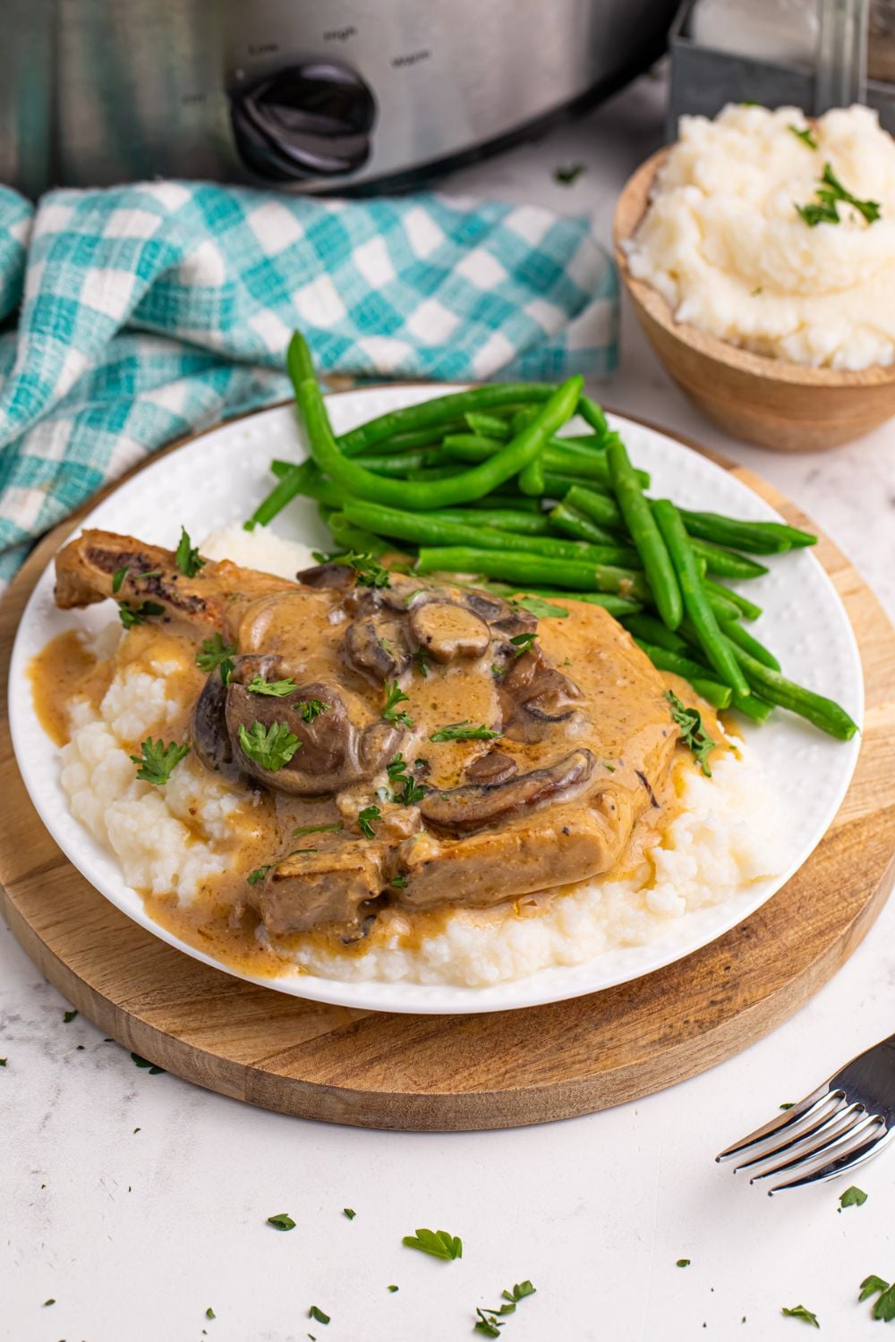 mashed potatoes on a plate with pork chop and gravy and green beans to the side with a crockpot behind it on the table 