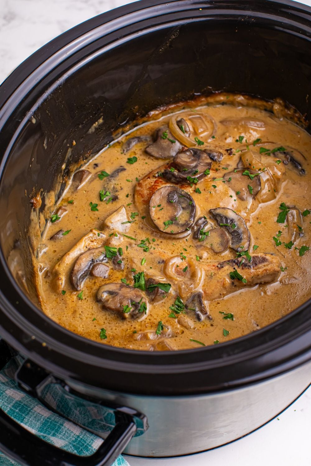 tilted black crockpot on table with pork chops cooked inside in a thick gravy with sauted mushroom and onion 
