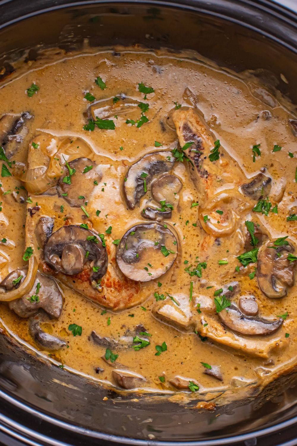 looking down to a crockpot on table with pork chops and gravy with mushrooms and fresh parsley on top