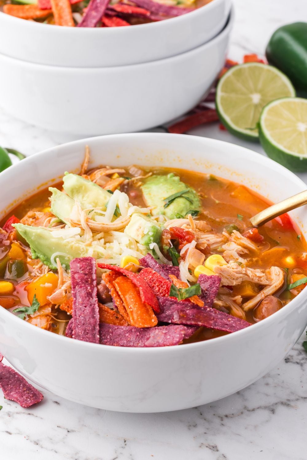 white bowl on table full of Southwestern Chicken Soup topped with avocado, shredded cheese, tortilla chips and more with more bowls stacked in the back of table with limes on the table