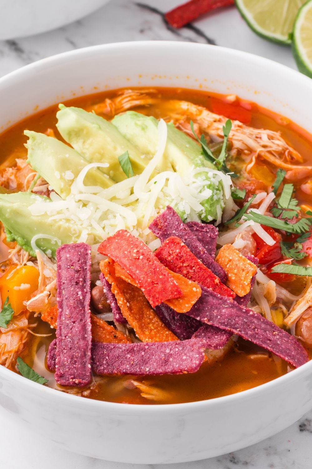 close up look of white bowl full of Southwestern Chicken Soup topped with tortilla strips, shredded cheese and sliced avocado