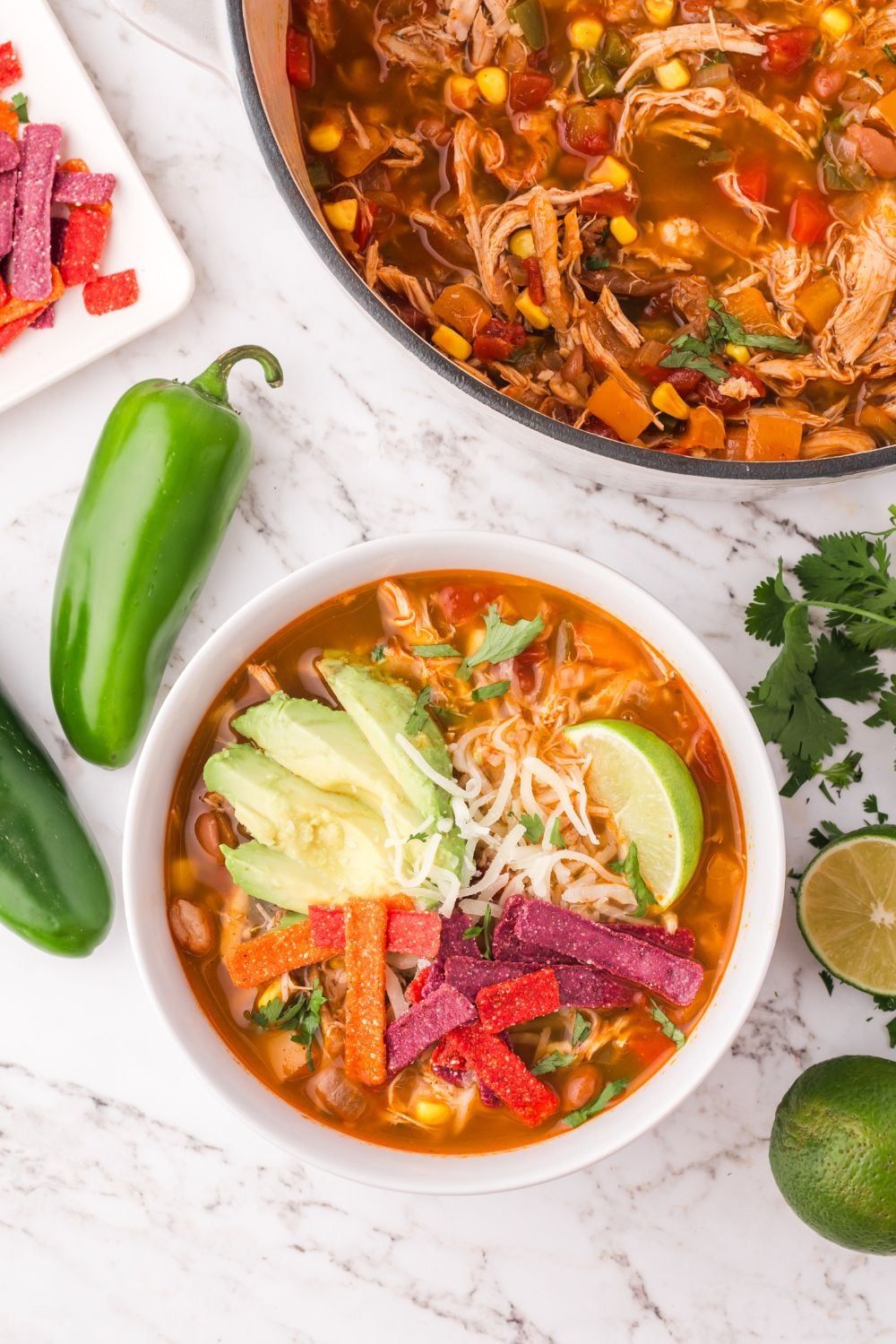 Southwestern Chicken Soup in a white bowl on the table and a pot farther back on the table topped with avocado, chips, cheese, and whole jalapenos on table