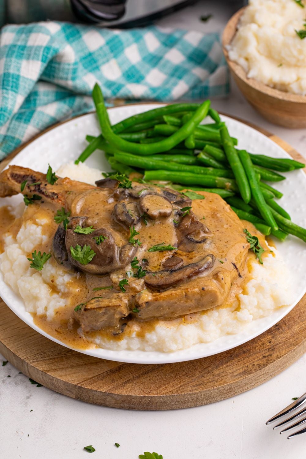 mashed potatoes on a plate with pork chop and gravy and green beans to the side with a crockpot behind it on the table