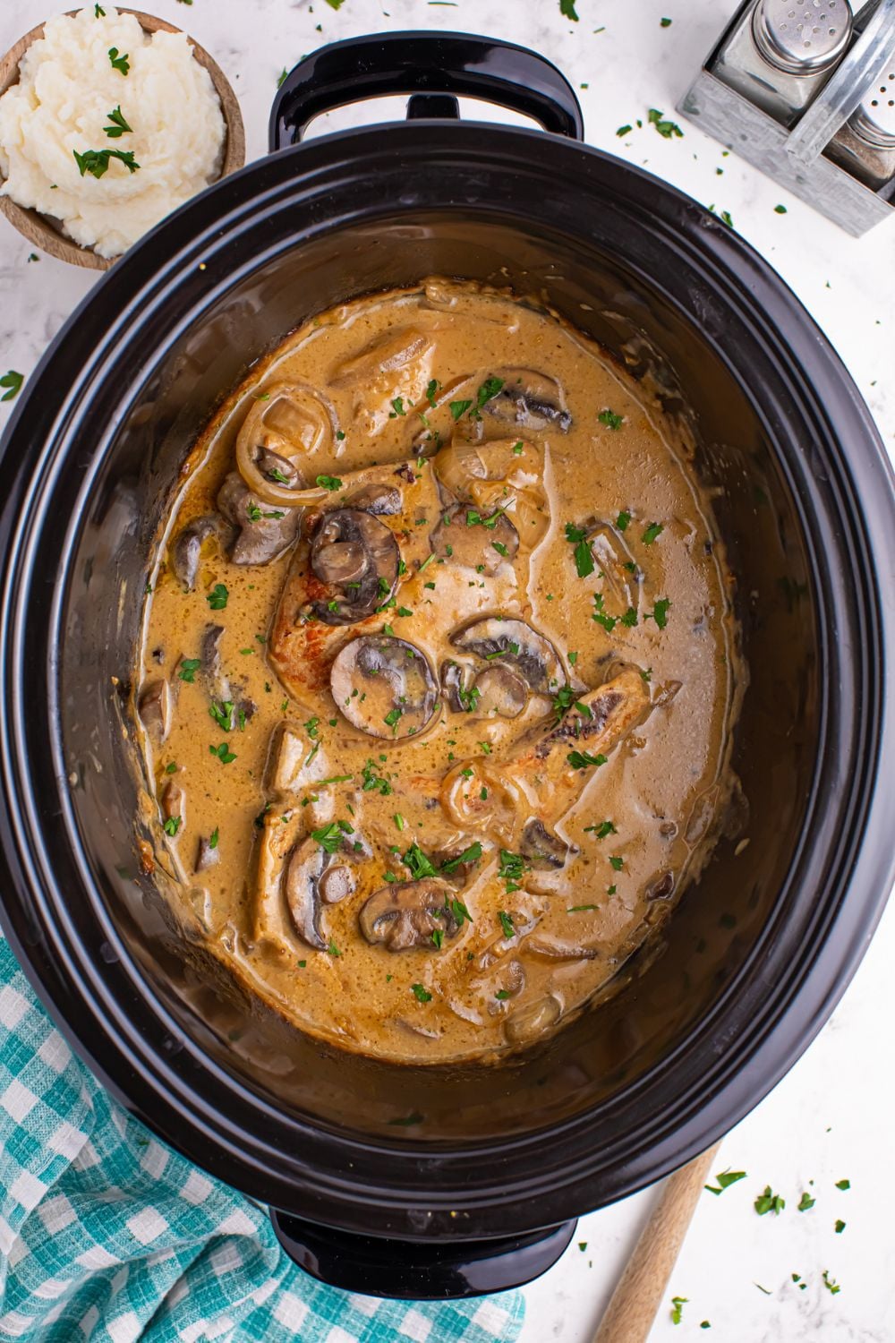 looking down at crockpot on table with a pork chop and gravy with parsley sprinkled on the top of it and a wooden spoon to the side 
