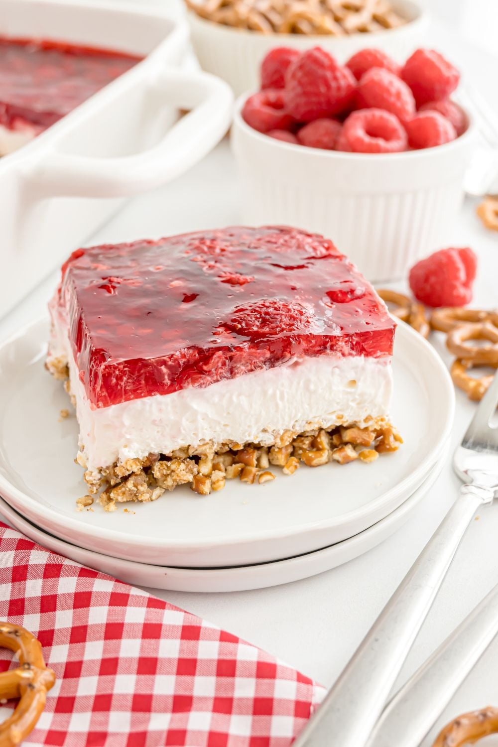 plate with a slice of Raspberry Pretzel Salad an then a bowl of raspberries to the side, and forks to the side