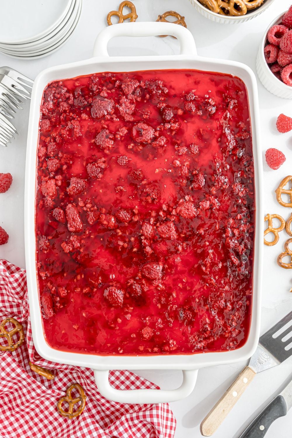 assembled Raspberry Jello salad with pretzels in a casserole dish on table with fruit and pretzels around the pan