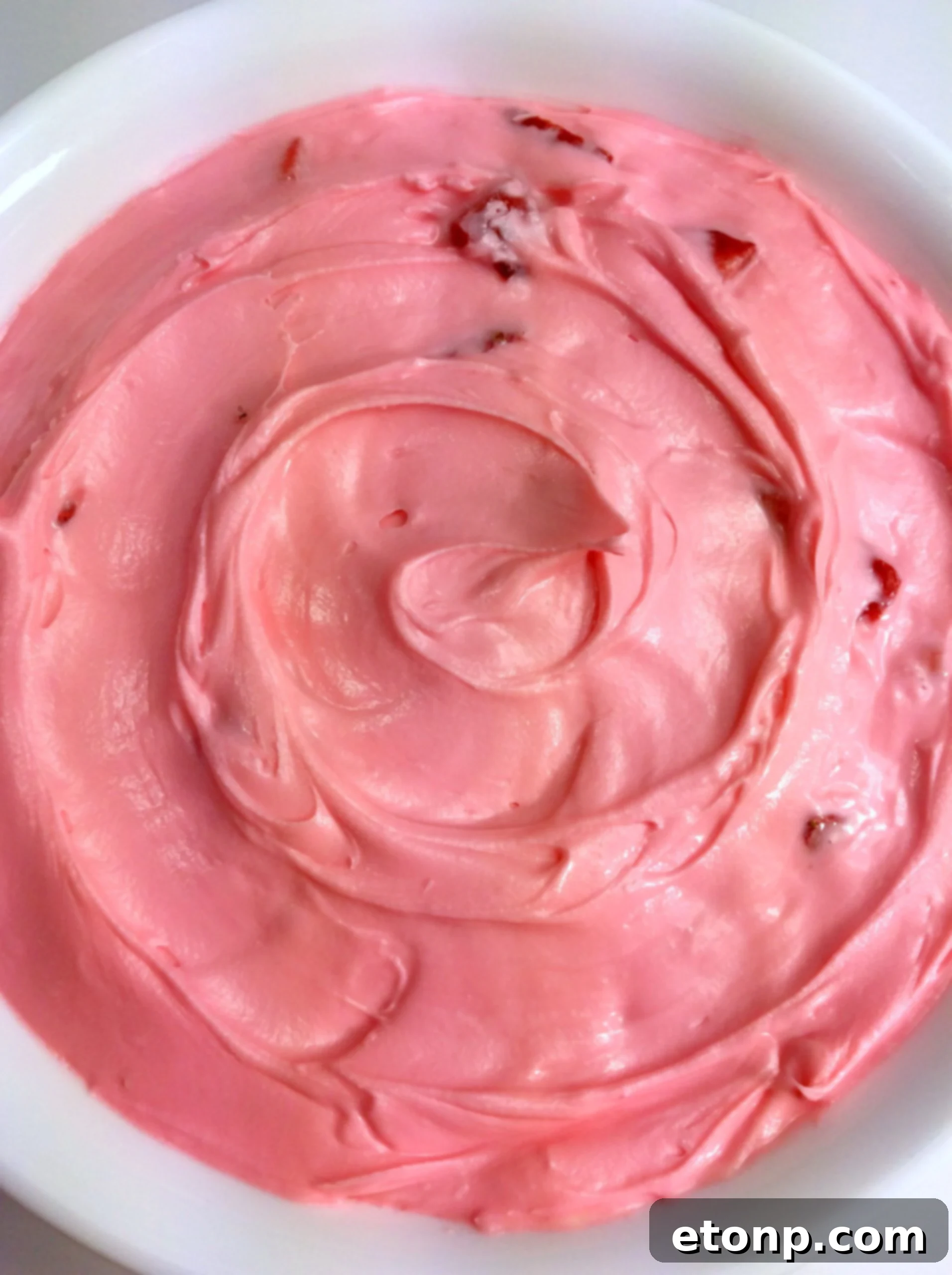 A close-up shot of a small bowl of strawberry buttercream frosting with a spoon.