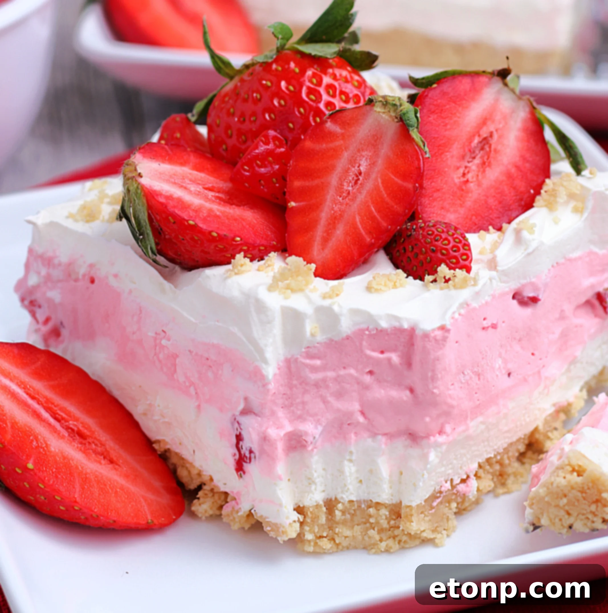 A slice of Strawberry Cheesecake Lasagna with a golden cookie crust, creamy cheesecake layer, vibrant strawberry gelatin, and a light whipped topping, garnished with fresh strawberries.